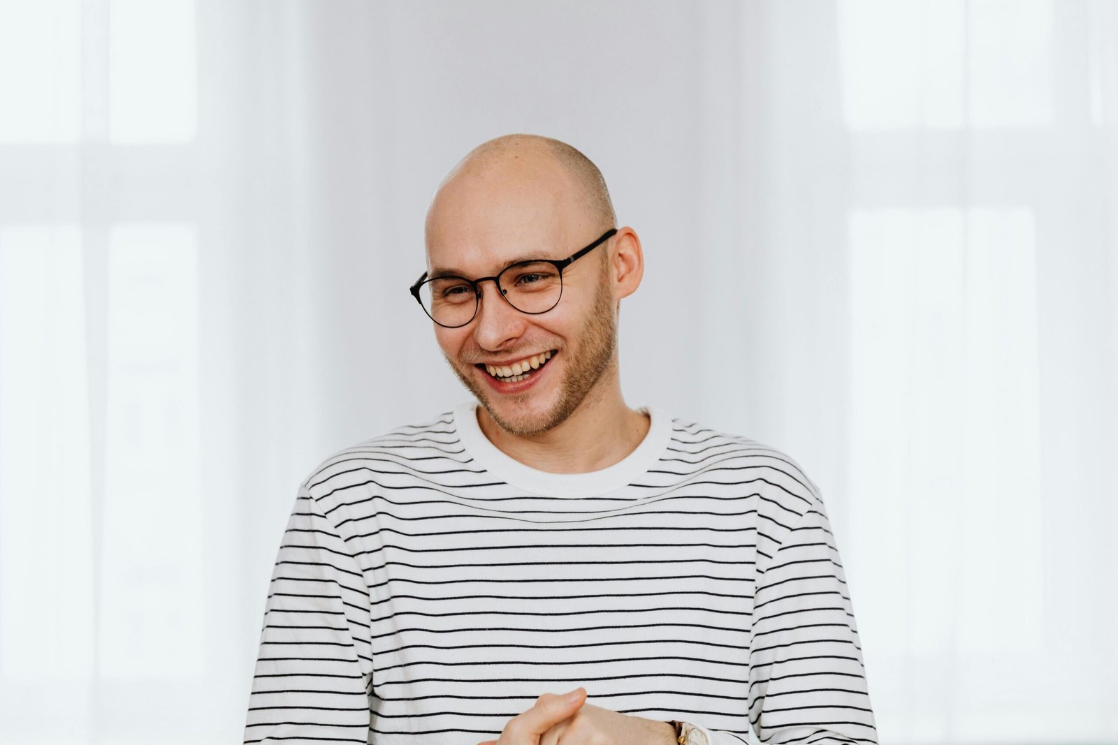 A bald man with glasses and a joyful smile in a striped shirt indoors.